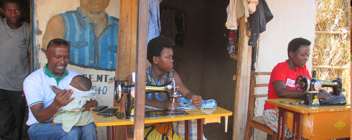 Two women sit at sewing machines whilst a man holds a baby outside a building