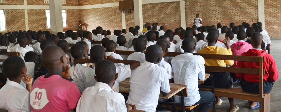 students in white shirts sit on benches listening to a lecture